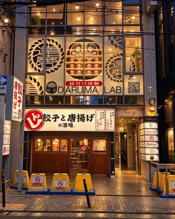 The DARUMA LAB. storefront at night in Nishi-Shinsaibashi, Osaka, with lit Daruma graphics on the 2nd floor